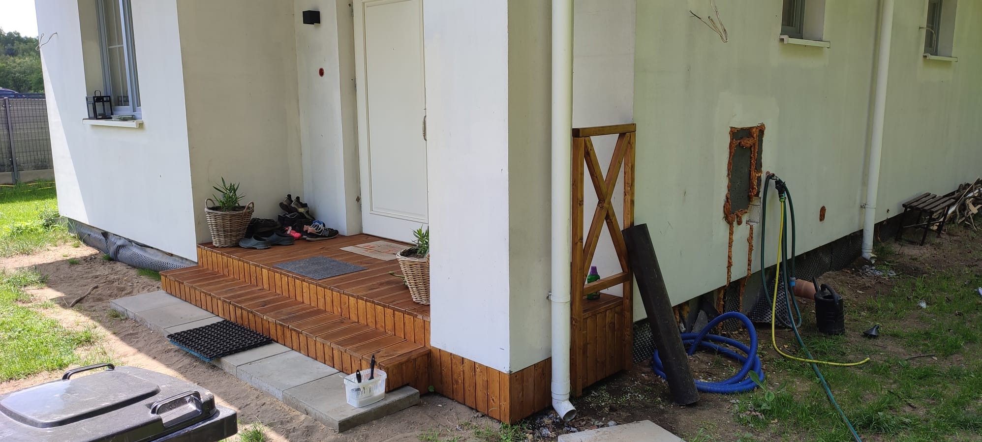 Drewniane schody wejściowe do domu z roślinami w doniczkach, obok widoczna drewniana konstrukcja. Na elewacji ślady po naprawach i węże ogrodowe.