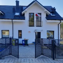 Otwarta, nowoczesna brama posesyjna z poziomych profili stalowych przed nowym, białym domem z czarną dachówką i flagą Polski na balkonie.