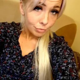 Portret kobiety z blond włosami i kwiecistą bluzką, siedzącej na beżowym fotelu, z lekkim uśmiechem i spojrzeniem w obiektyw.