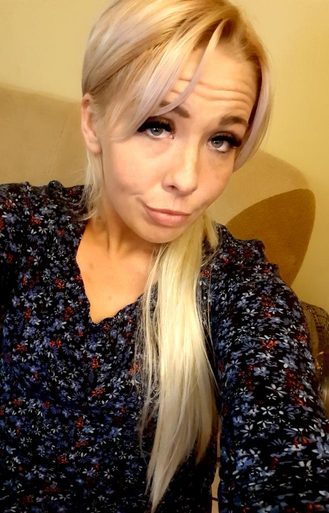 Portret kobiety z blond włosami i kwiecistą bluzką, siedzącej na beżowym fotelu, z lekkim uśmiechem i spojrzeniem w obiektyw.