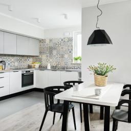 LMBUD sp. z o. o. - Jasna kuchnia z białymi szafkami, wzorzystą mozaiką nad blatem i czarnymi krzesłami przy białym stole, oświetlona nowoczesną lampą wiszącą.
