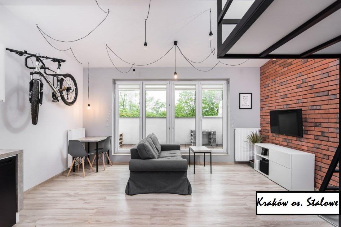 Jasne wnętrze pokoju dziennego z szarą sofą, ścianą z czerwonej cegły i rowerem zawieszonym na ścianie, oświetlone wiszącymi żarówkami na kablach.