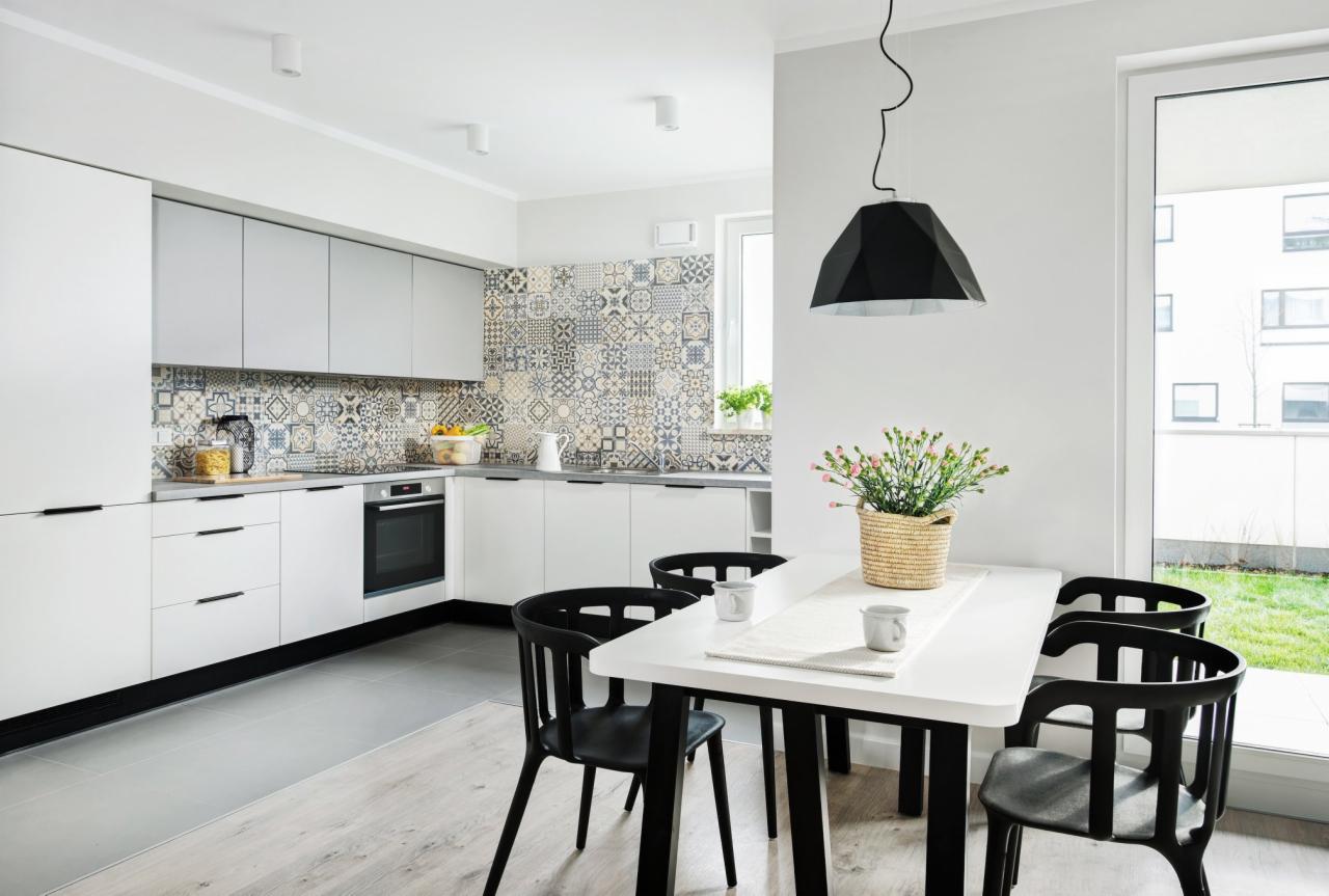 Jasna kuchnia z białymi szafkami, wzorzystą mozaiką nad blatem i czarnymi krzesłami przy białym stole, oświetlona nowoczesną lampą wiszącą.