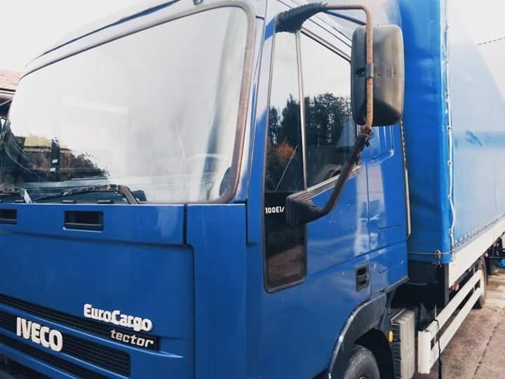 Niebieski samochód ciężarowy marki Iveco EuroCargo z plandeką, widoczny z boku, z lusterkiem wstecznym na wysięgniku i oznaczeniem modelu 100E1/