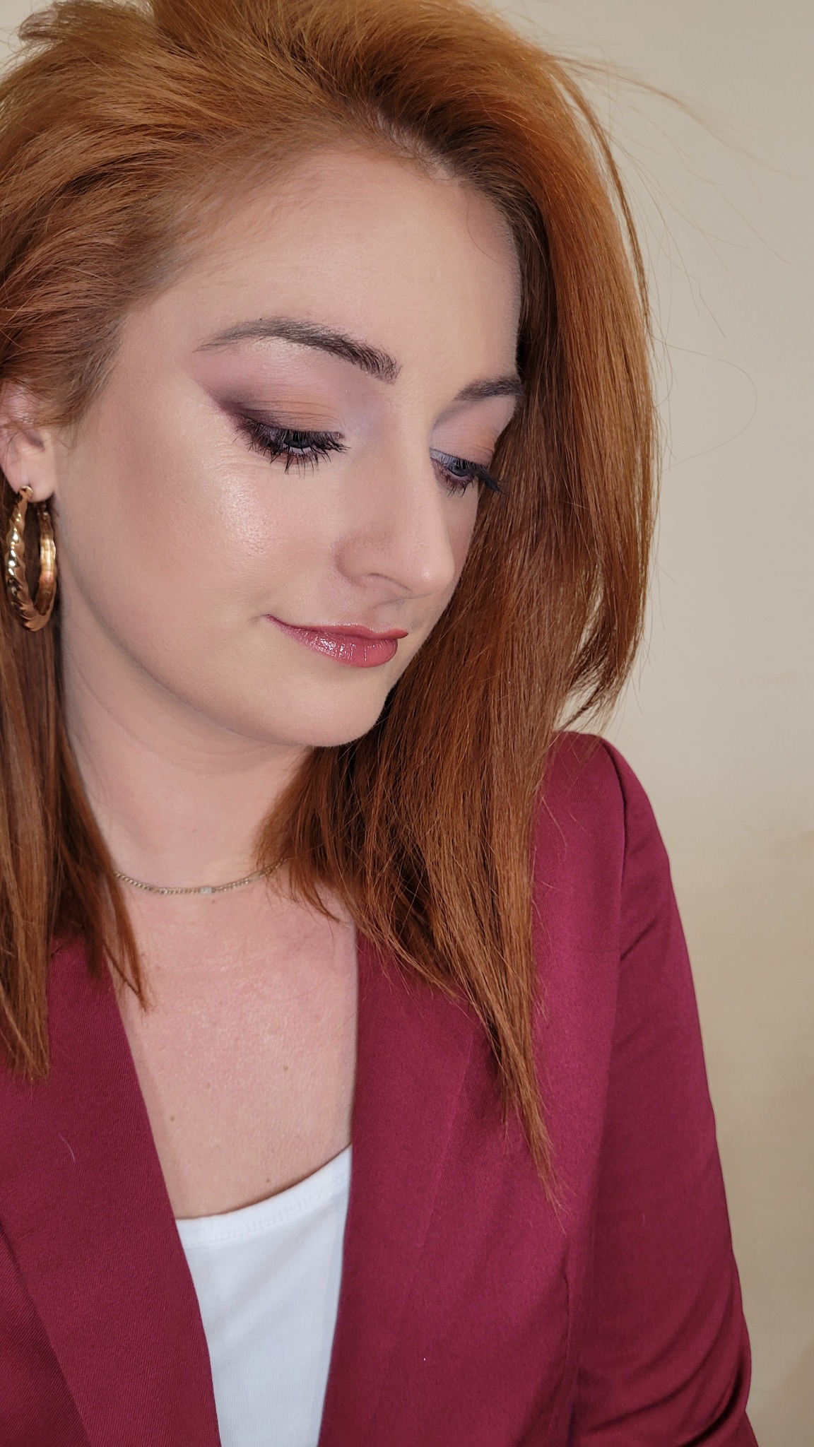 Portret kobiety z rudymi włosami, delikatny makijaż oczu w odcieniach fioletu i błyszczące usta, ubrana w bordową marynarkę i złote kolczyki.