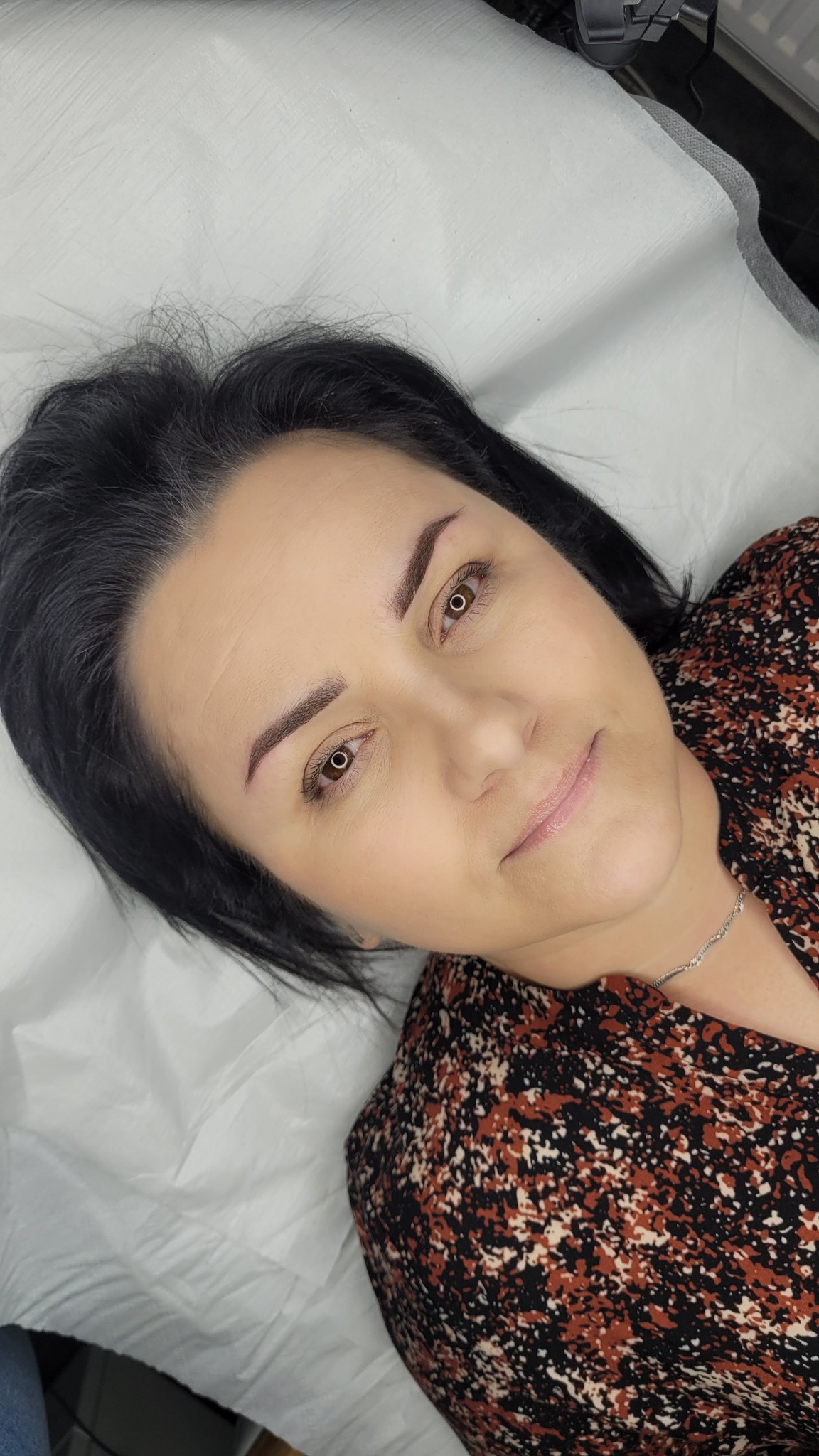 Kobieta leżąca na białym prześcieradle, po zabiegu pigmentacji brwi metodą pudrową, widoczne świeżo wykonane brwi o naturalnym kształcie i kolorze.