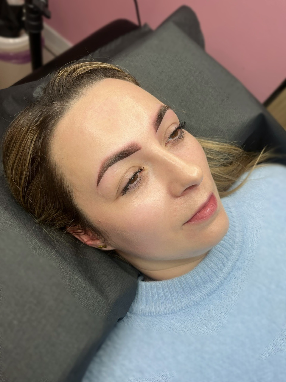 Kobieta leżąca na poduszce z widocznymi brwiami po zabiegu microbladingu, delikatny makijaż permanentny brwi, jasna cera, błękitny sweter.