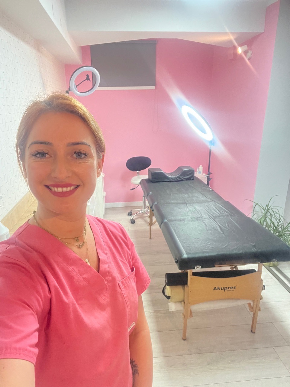 Uśmiechnięta kosmetyczka w różowym uniformie na tle gabinetu z czarnym leżanką do zabiegów i lampami pierścieniowymi