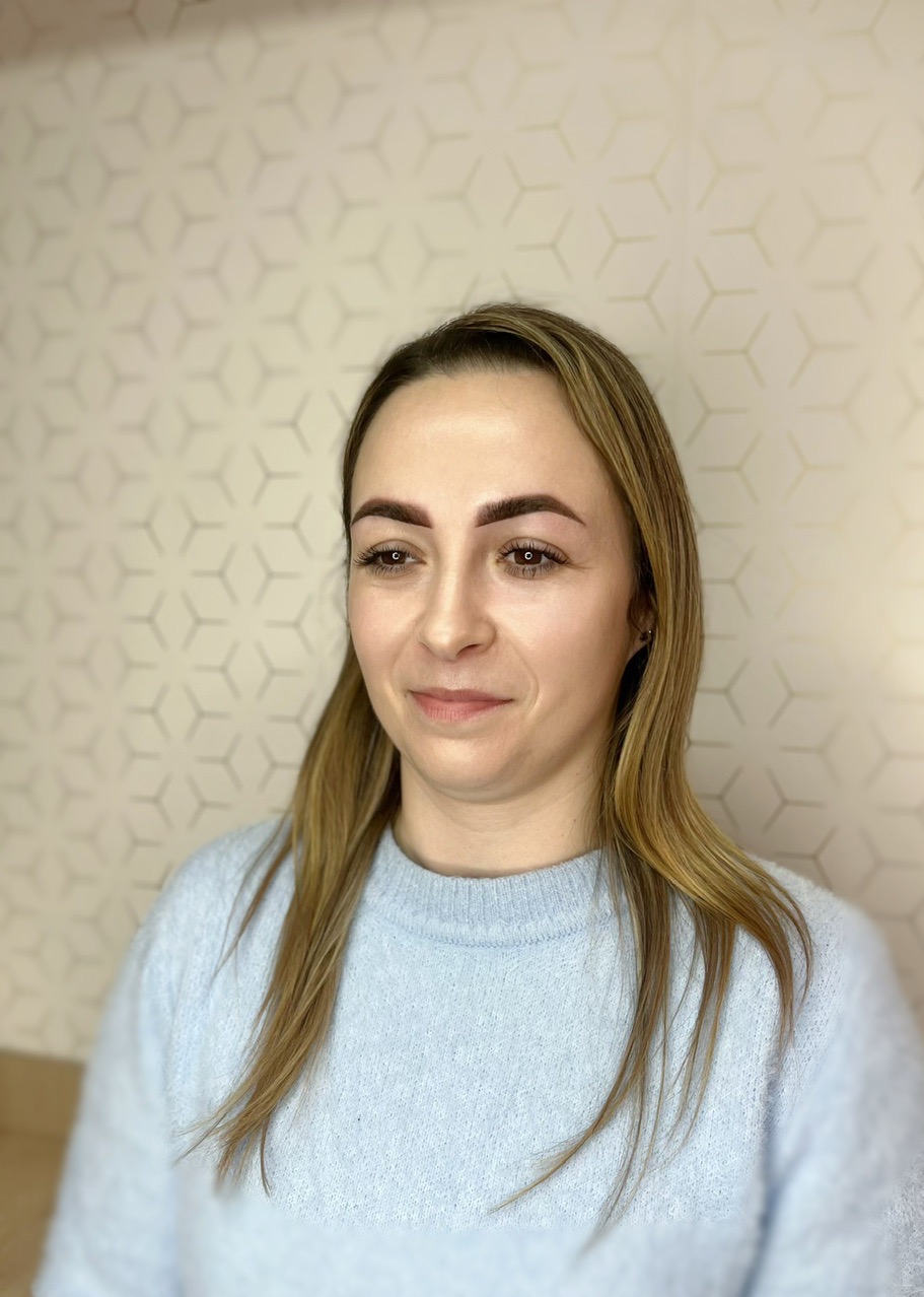 Portret kobiety z brązowymi brwiami po zabiegu makijażu permanentnego, na tle ściany z geometrycznym wzorem.