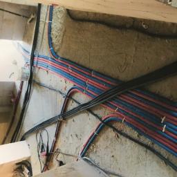 Stasbud - Świeżo poprowadzone rury instalacji wodnej i elektrycznej w ścianie podczas remontu, rury czerwone i niebieskie ułożone równolegle, czarne kable elektryczne w peszlach, surowy stan wykończenia.