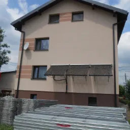 Elewacja domu z zamontowanymi kolektorami słonecznymi, widoczne stosy metalowych rusztowań budowlanych na trawniku.