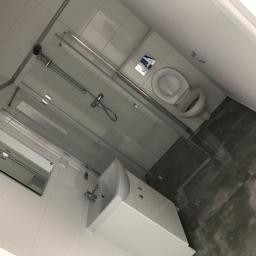 Stasbud - Nowoczesna łazienka po remoncie: białe płytki, kabina prysznicowa ze szklanymi drzwiami, umywalka z szafką i toaleta z podtynkową spłuczką, szare płytki podłogowe imitujące beton.