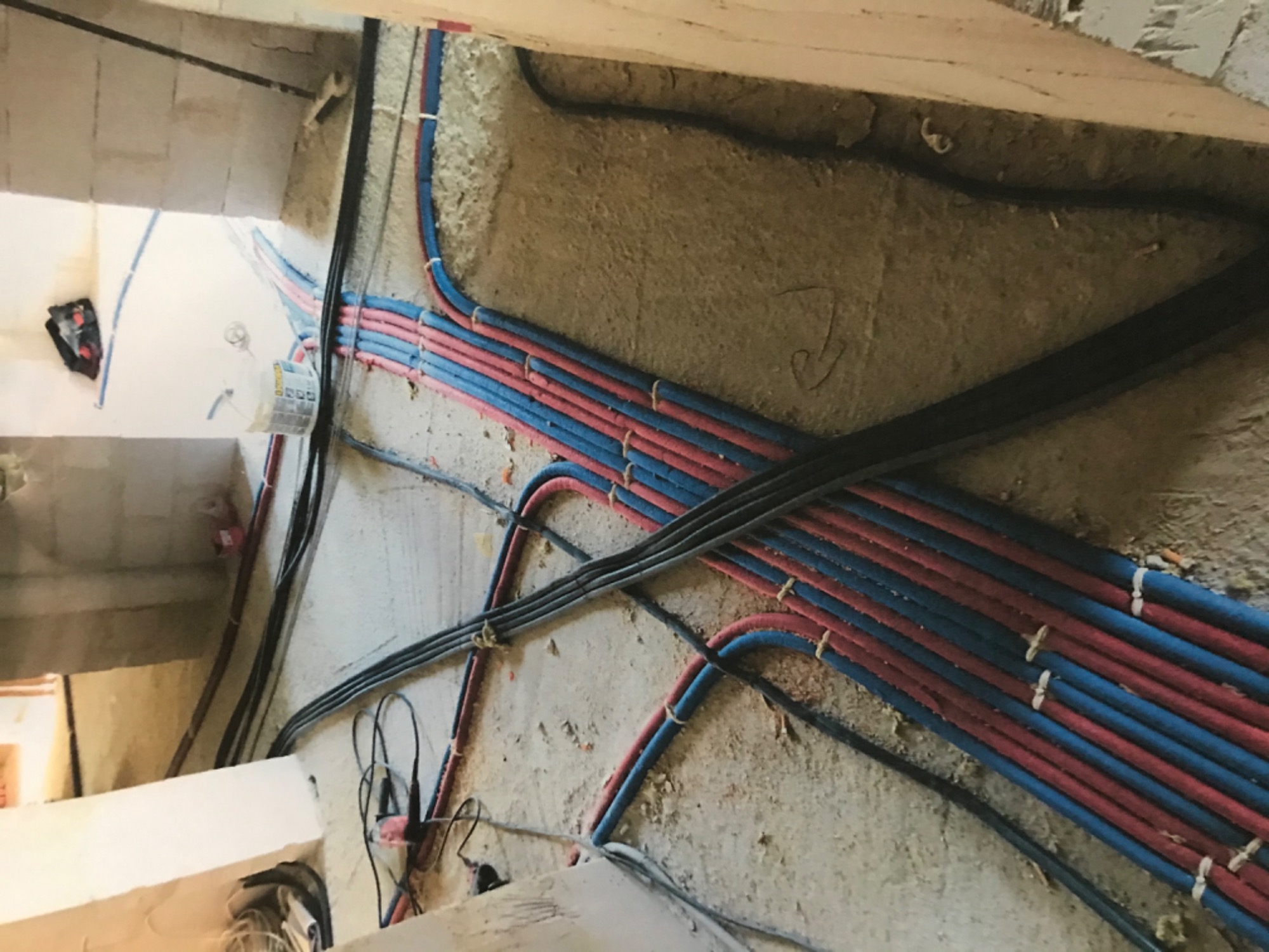 Świeżo poprowadzone rury instalacji wodnej i elektrycznej w ścianie podczas remontu, rury czerwone i niebieskie ułożone równolegle, czarne kable elektryczne w peszlach, surowy stan wykończenia.