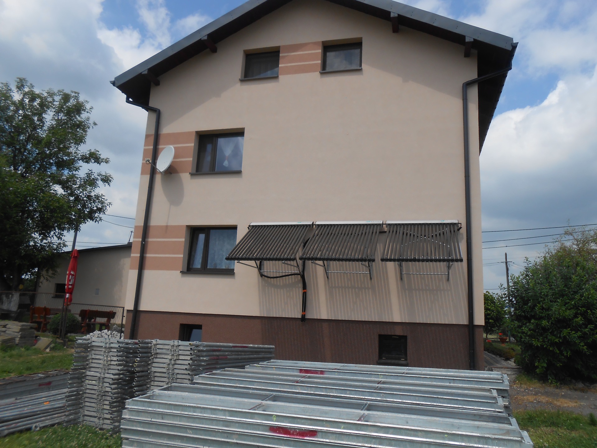 Elewacja domu z zamontowanymi kolektorami słonecznymi, widoczne stosy metalowych rusztowań budowlanych na trawniku.