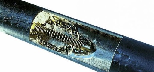 Przekrój rury kanalizacyjnej z widocznym wewnątrz sprężynowym narzędziem do udrażniania, otoczonym fragmentami zanieczyszczeń.