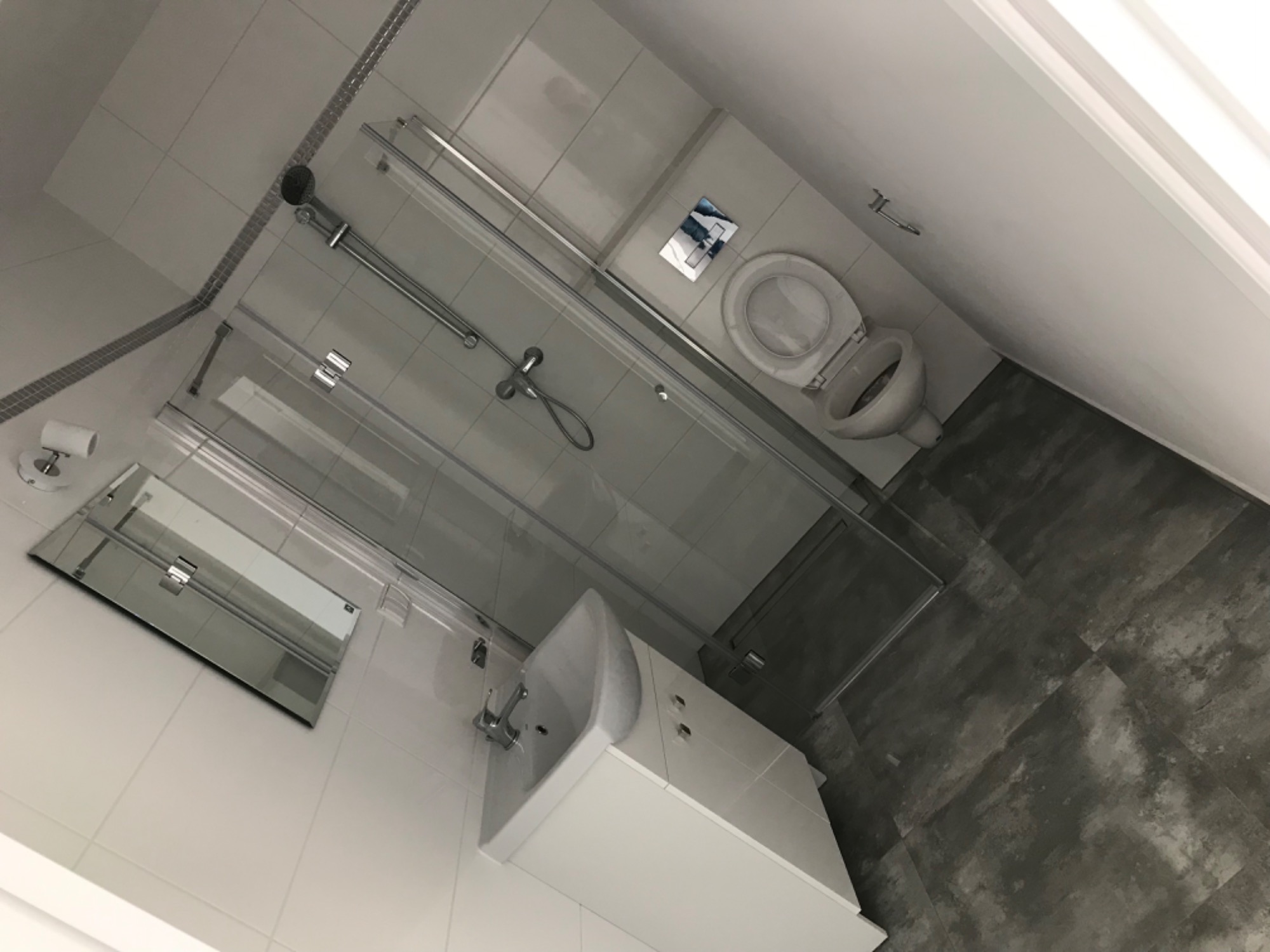 Nowoczesna łazienka po remoncie: białe płytki, kabina prysznicowa ze szklanymi drzwiami, umywalka z szafką i toaleta z podtynkową spłuczką, szare płytki podłogowe imitujące beton.