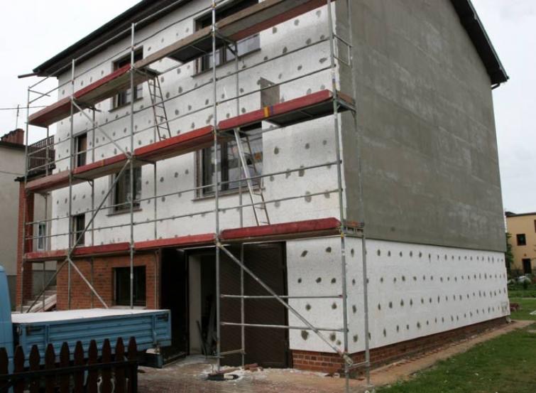 Ocieplanie elewacji dwukondygnacyjnego domu jednorodzinnego, widoczne rusztowania i fragment otynkowanej ściany szczytowej.