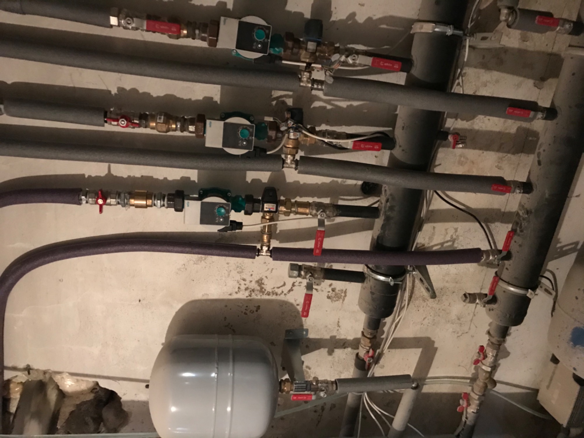 Rozbudowana instalacja centralnego ogrzewania z rurami, zaworami, pompami obiegowymi i zbiornikiem wyrównawczym, widok z dołu na tle szarej ściany.