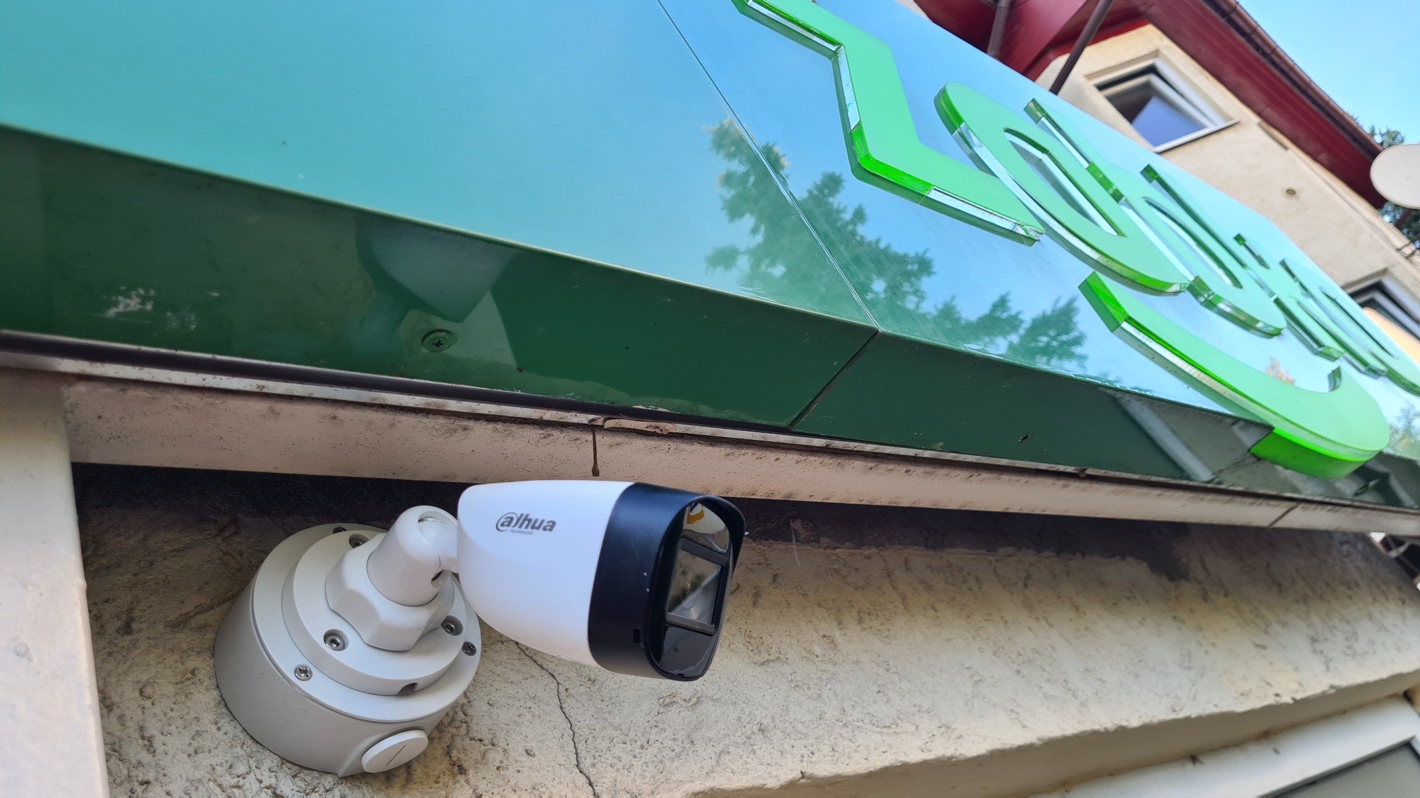 Biała kamera monitoringu marki Dahua zamontowana na szarej ścianie budynku pod zielonym szyldem sklepu, widok z dołu.