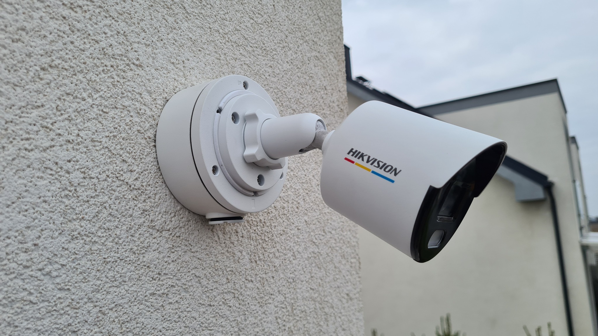 Biała kamera monitoringu marki Hikvision zamontowana na chropowatej, jasnej elewacji budynku jednorodzinnego, widoczna z bliskiej perspektywy.