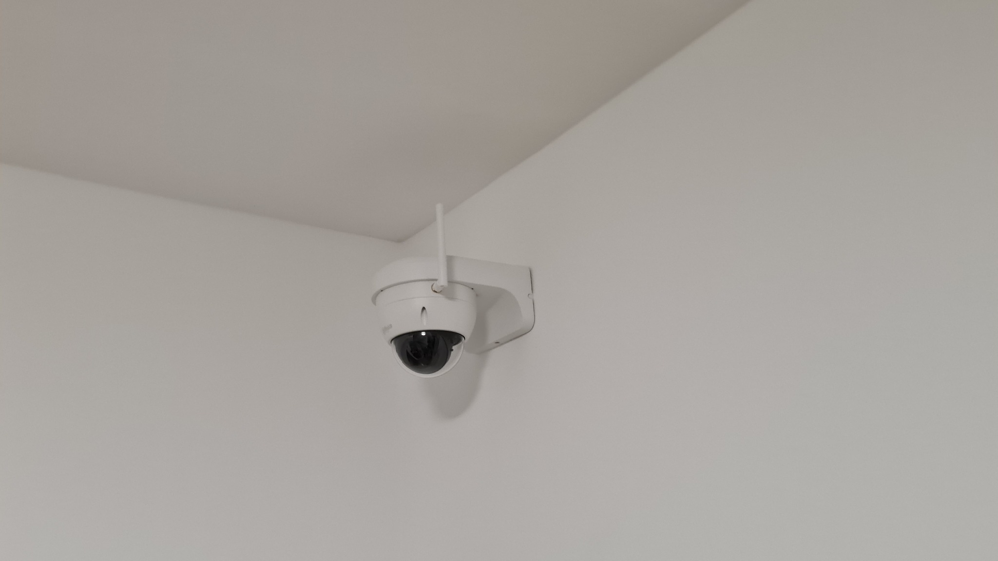 Biała kamera kopułkowa z anteną, zamontowana na białej ścianie w rogu pomieszczenia, widok z dołu.