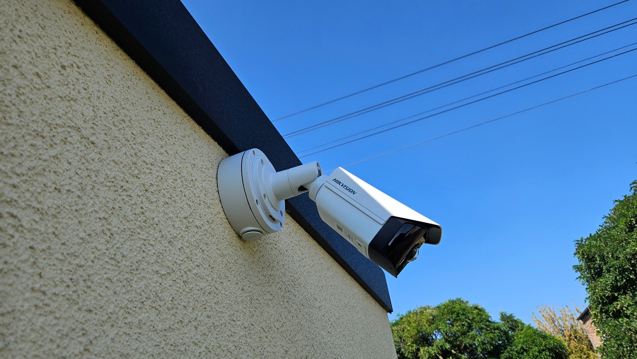 Biała kamera Hikvision zamontowana na elewacji budynku o jasnej, chropowatej strukturze, z widocznymi przewodami elektrycznymi na tle błękitnego nieba.