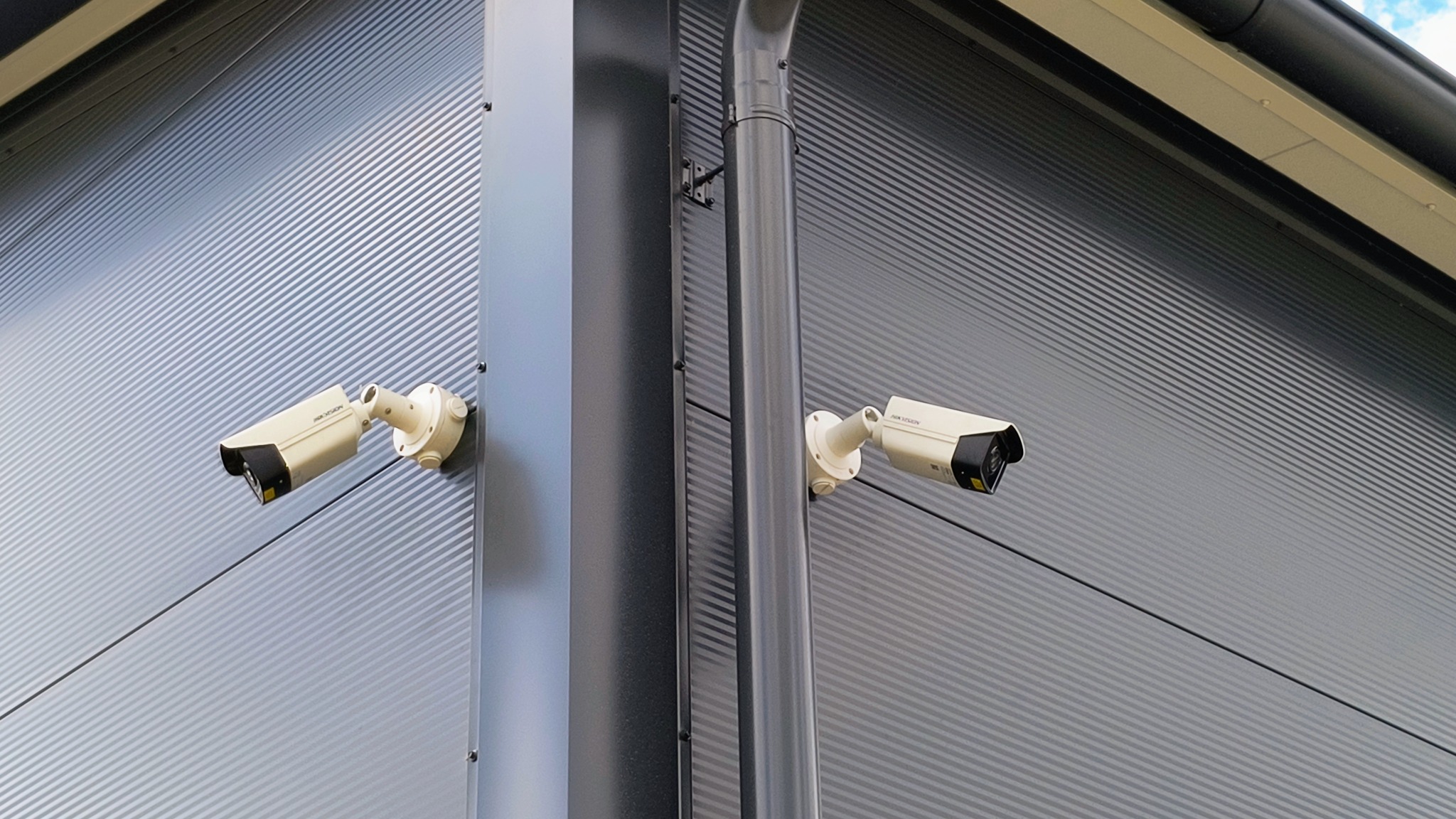 Dwie kamery przemysłowe zamontowane na szarej, metalowej elewacji budynku, jedna przy narożniku, druga obok rury spustowej.