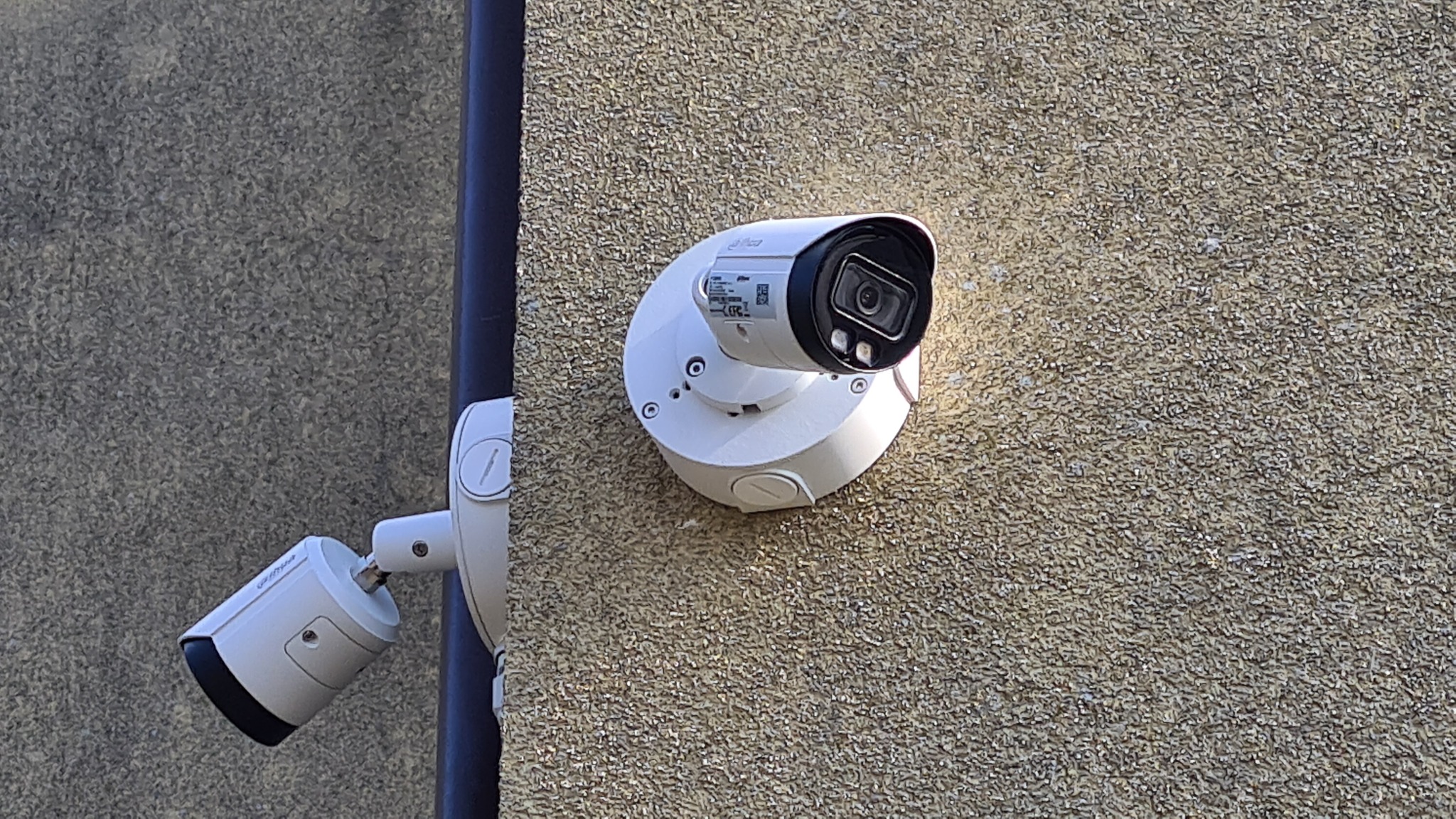 Dwie kamery IP na elewacji budynku o grubej, ziarnistej fakturze. Jedna kamera kopułkowa zamontowana na okrągłej podstawie, druga cylindryczna, z widocznym uchwytem montażowym.