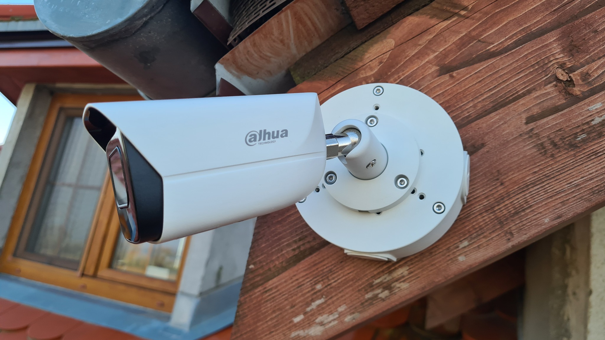 Biała kamera monitoringu Dahua zamontowana na drewnianej podbitce dachu z widocznym oknem w tle.