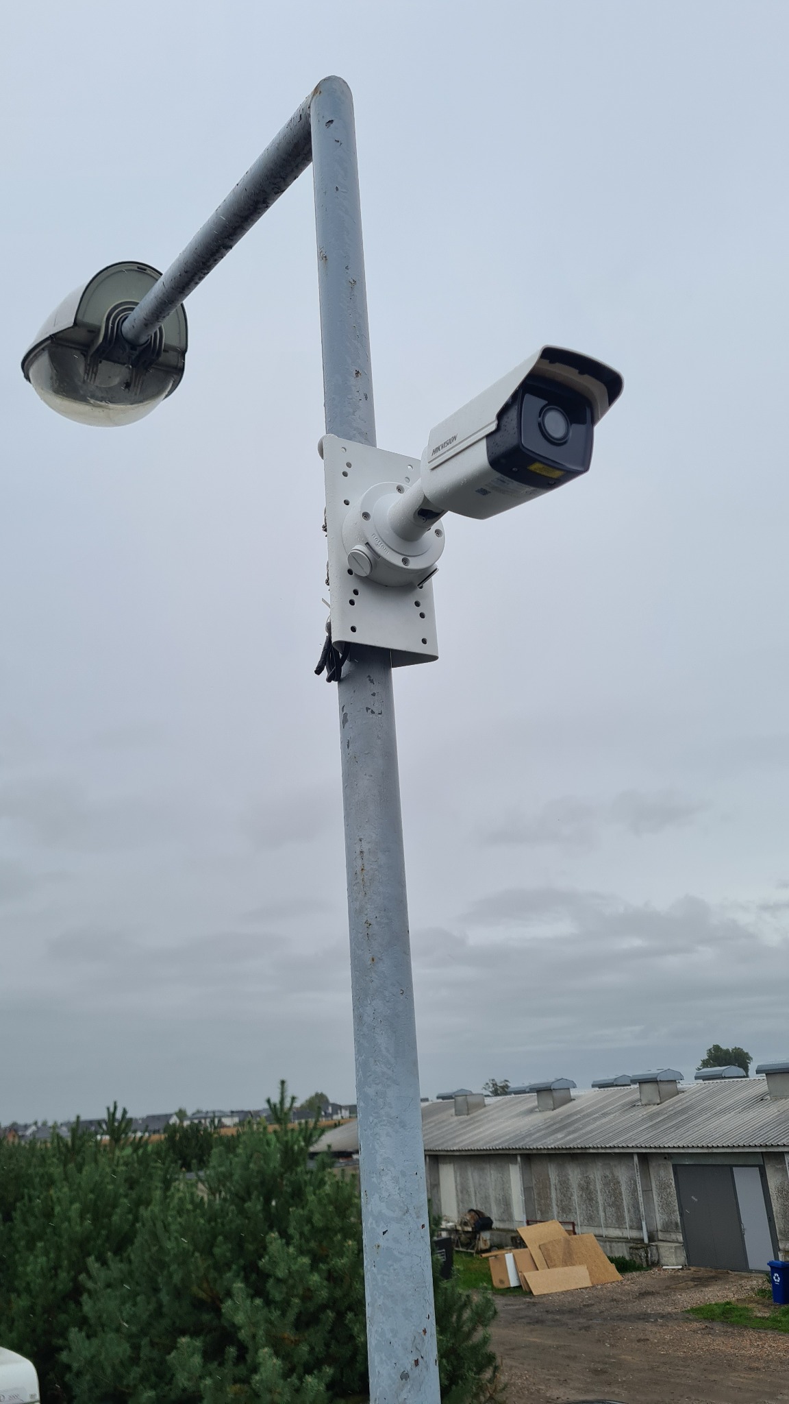 Kamera przemysłowa Hikvision zamontowana na metalowym słupie oświetleniowym na tle pochmurnego nieba i budynków gospodarczych.