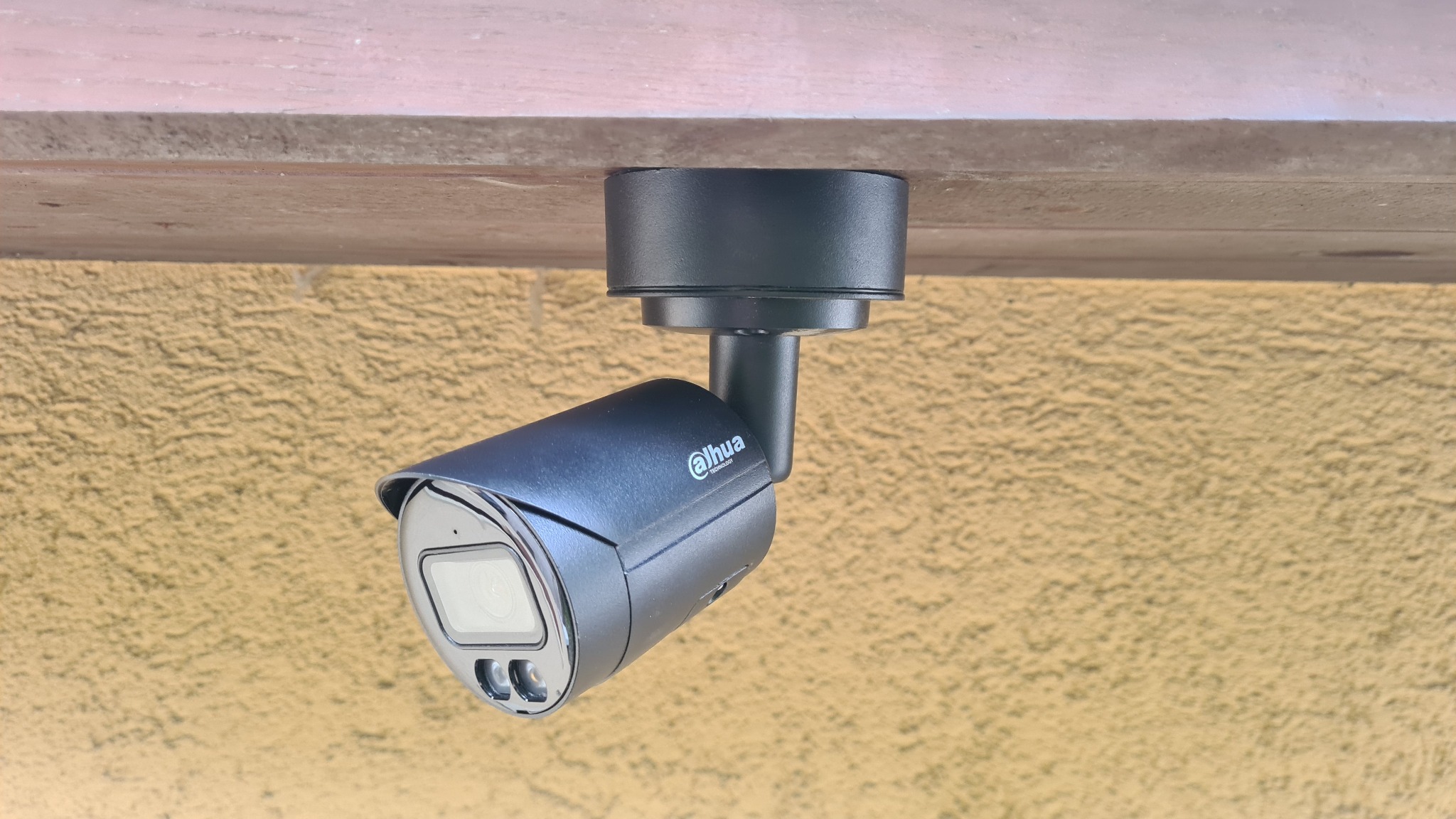 Czarna kamera monitoringu Dahua zamontowana pod drewnianym zadaszeniem, na tle żółtej, teksturowanej ściany.