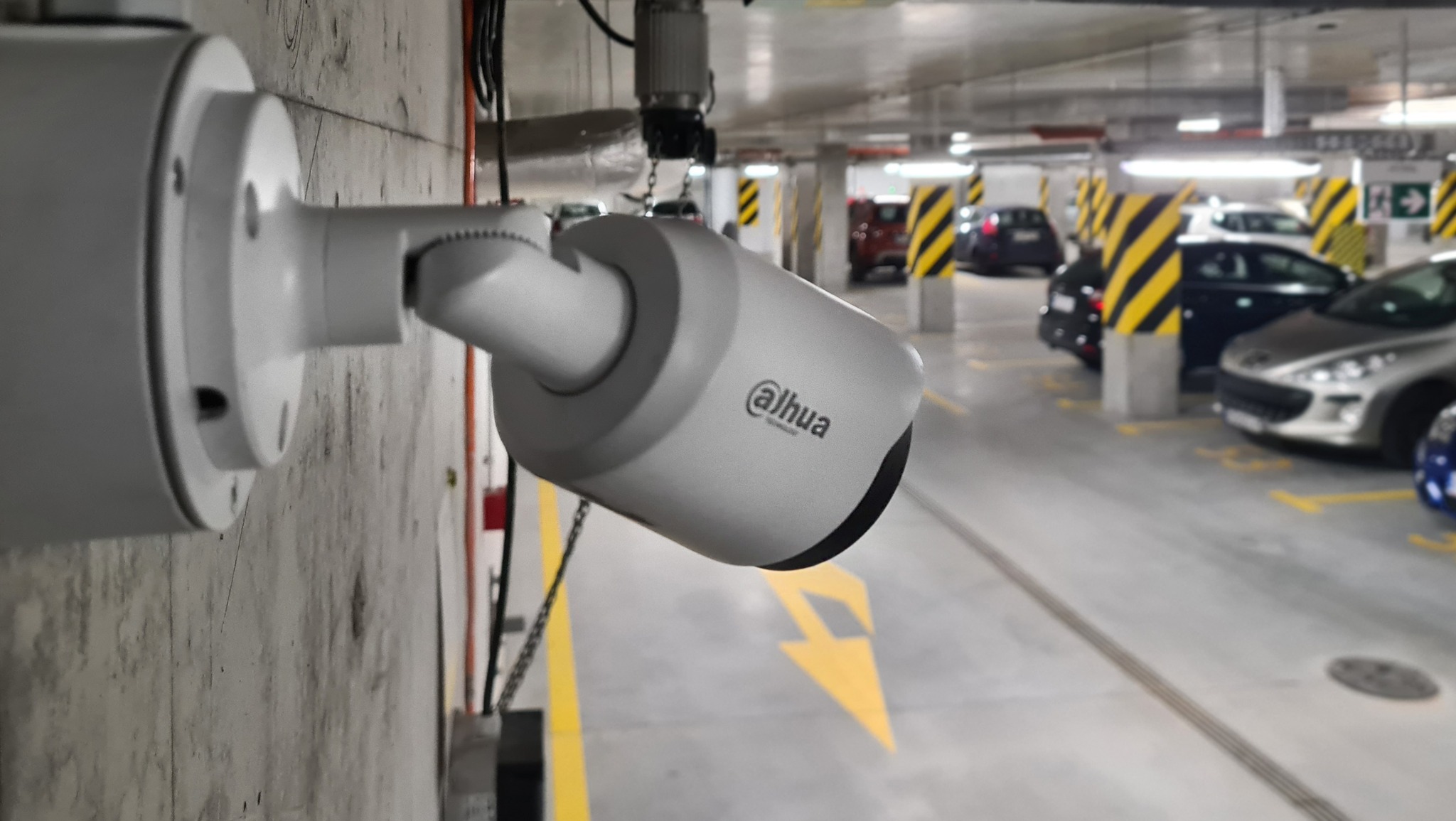 Kamera przemysłowa marki Dahua zamontowana na betonowej ścianie parkingu podziemnego, widoczne żółte pasy i zaparkowane samochody w tle.