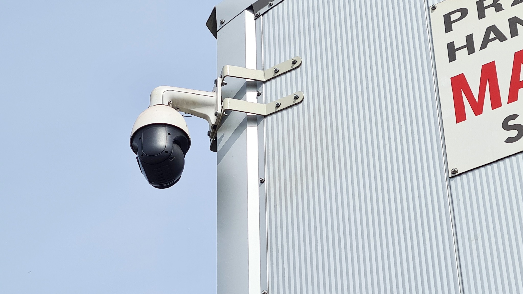 Zewnętrzna kamera obrotowa PTZ zamontowana na metalowej konstrukcji budynku z widocznym fragmentem szyldu reklamowego.