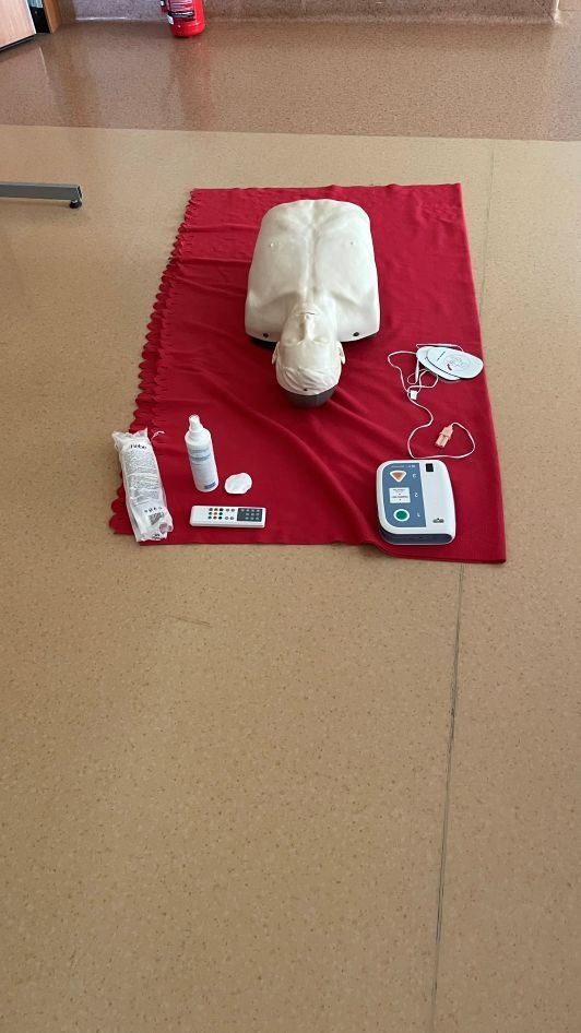 Manekin do resuscytacji na czerwonym materiale, obok defibrylator, środki dezynfekcyjne i akcesoria. Sprzęt do kursu pierwszej pomocy.