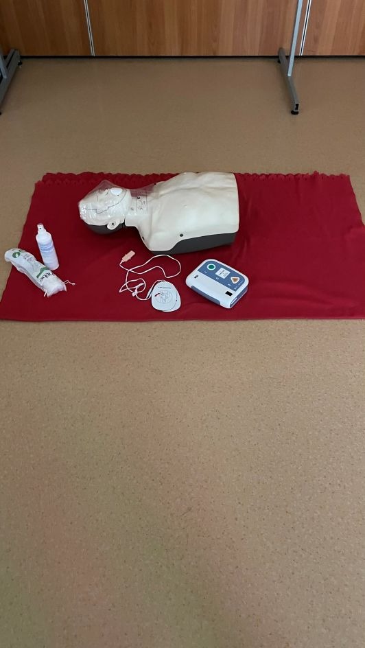 Manekin do resuscytacji na czerwonym podkładzie, obok defibrylator, płyn dezynfekujący i maska. Sprzęt do ćwiczeń z zakresu pierwszej pomocy.