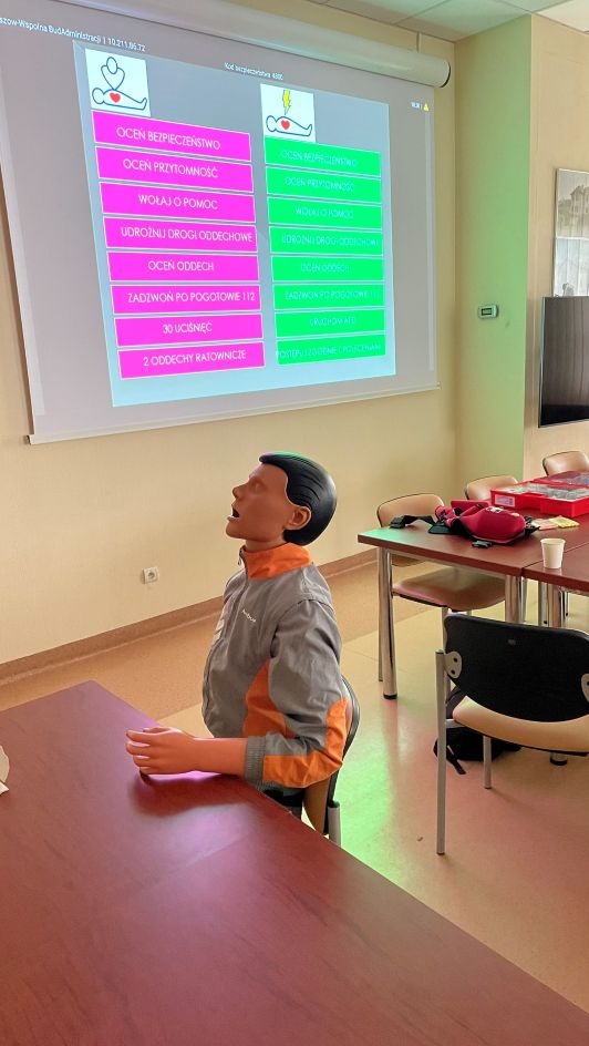 Manekin do resuscytacji na tle ekranu z instrukcjami pierwszej pomocy. Sala szkoleniowa z stołami i krzesłami. Szkolenie z udzielania pierwszej pomocy.