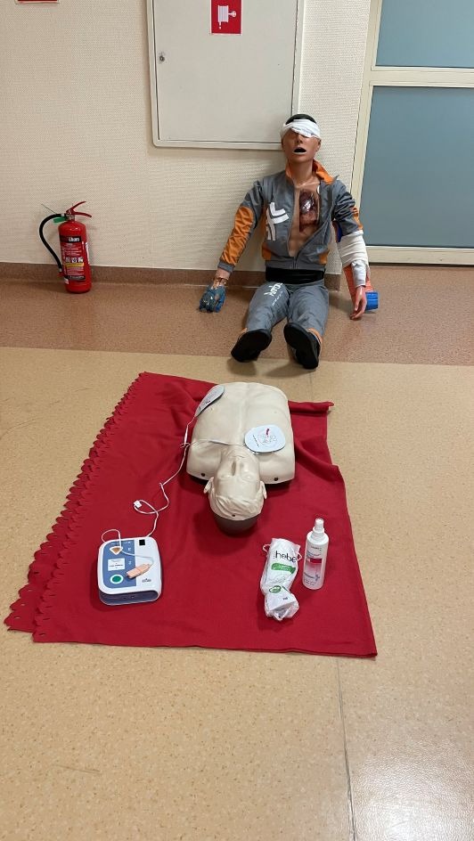 Manekin z opatrunkami i fantom na czerwonym materiale, obok defibrylator i środki higieniczne. W tle gaśnica i szafka hydrantowa. Scena z kursu pierwszej pomocy.