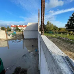 Wznoszenie ścian budynku z betonu komórkowego z widocznym zbrojeniem pionowym, na zalanym błotem placu budowy. W tle budynek mieszkalny i błękitne niebo.