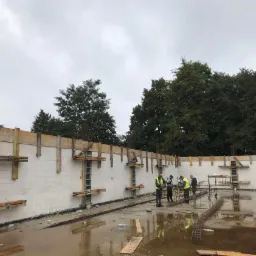 Budowa fundamentów domu murowanego: widoczne szalunki, zbrojenia i pracownicy w żółtych kamizelkach na zalanej wodą powierzchni.