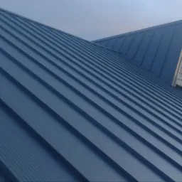 Nowy, blaszany dach w kolorze grafitowym z widocznymi rąbkami, fragment rusztowania i pracownik w oddali, w tle drzewa i pochmurne niebo.