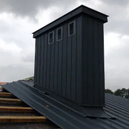 Szary komin wentylacyjny z trzema rzędami otworów na dachu z blachy trapezowej, w tle zachmurzone niebo i fragment krajobrazu.