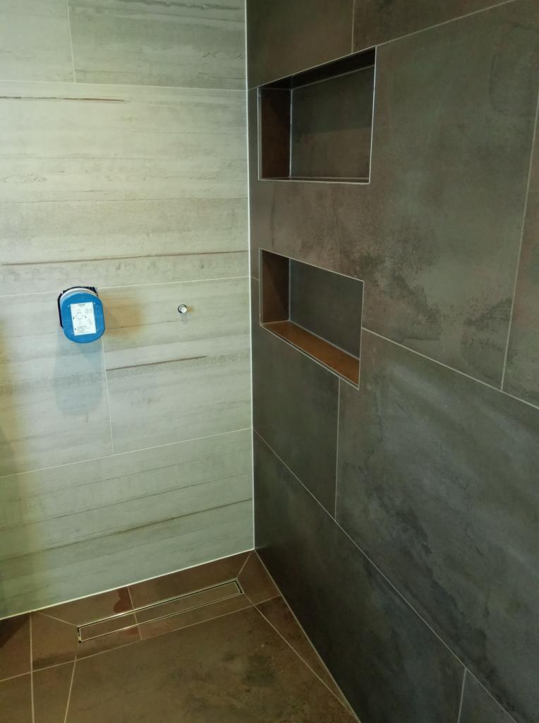 Wykończona łazienka z szarymi i beżowymi płytkami, wbudowanymi półkami i odpływem liniowym w podłodze.