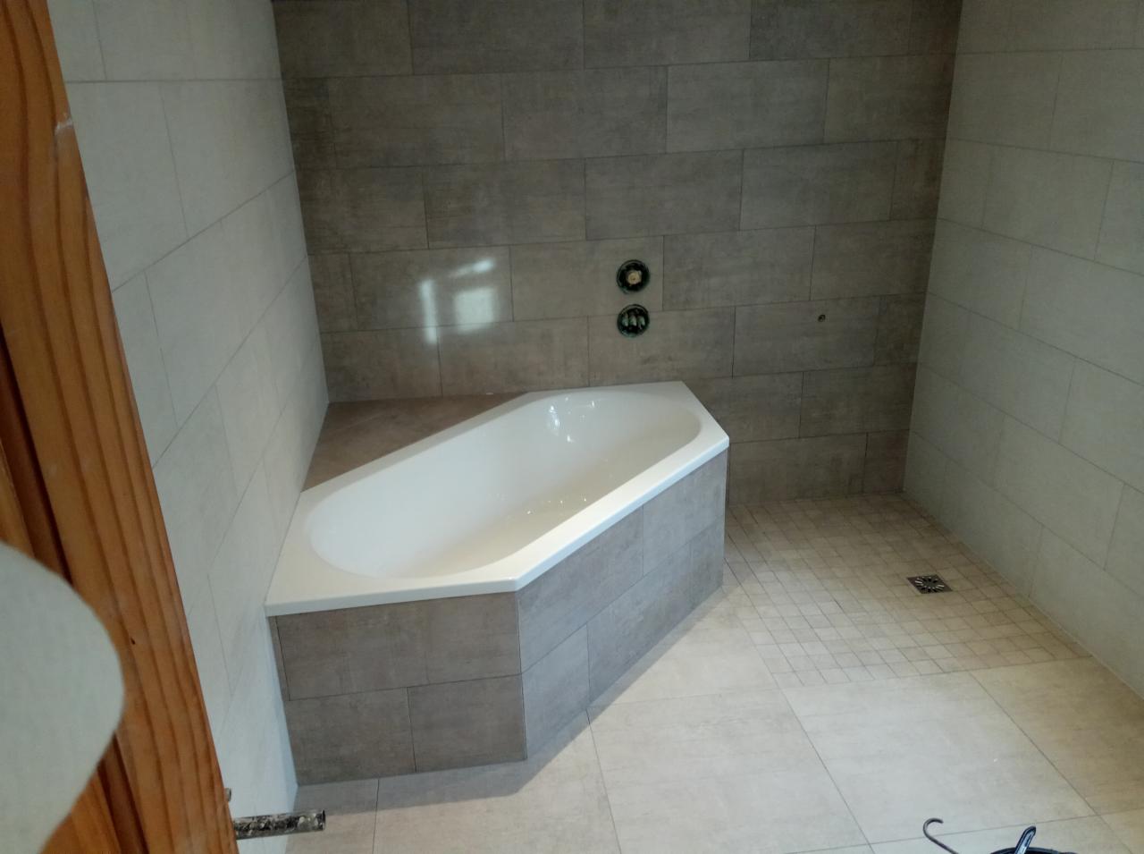 Narożna wanna obudowana płytkami, z widocznymi zamontowanymi bateriami i odpływem podłogowym, całość wyłożona płytkami w odcieniach szarości.