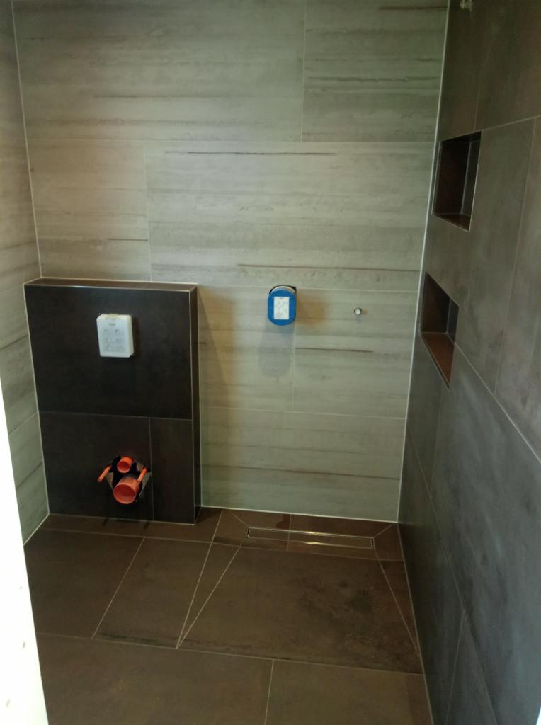 Wykończona łazienka z brązowymi i beżowymi płytkami imitującymi drewno, zabudowa WC z przyciskiem spłukującym, odpływ liniowy w podłodze i wnęki w ścianie.