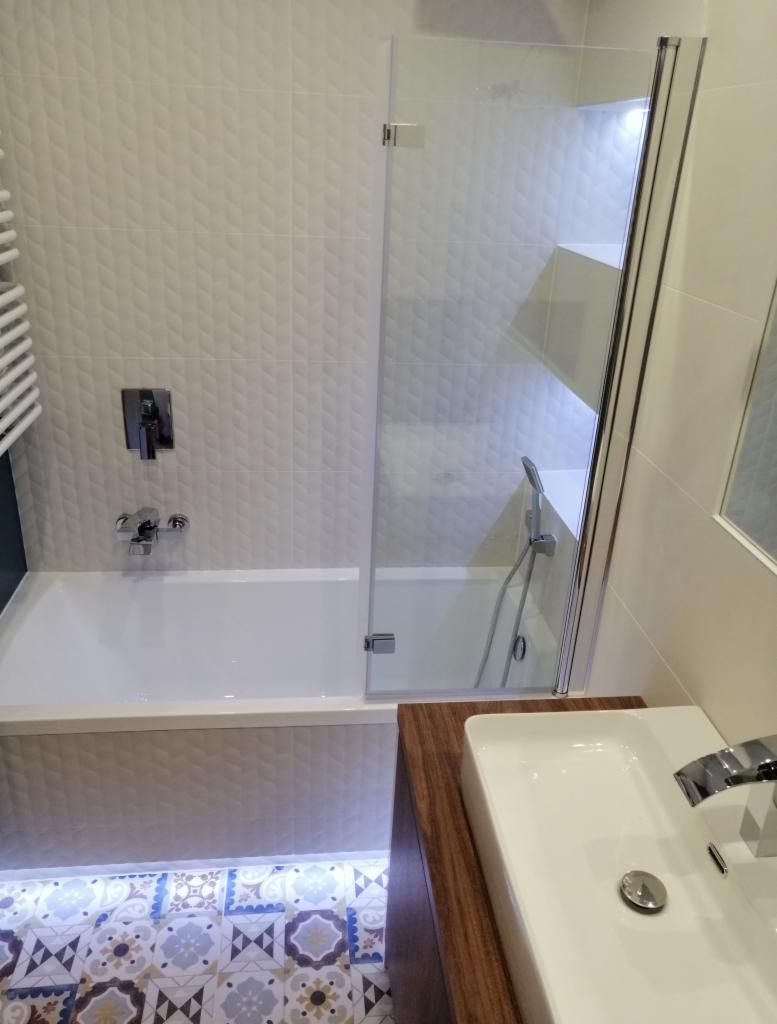 Wykończona łazienka z białą wanną obudowaną teksturowanymi płytkami, szklanym parawanem nawannowym, umywalką nablatową na drewnianej szafce i dekoracyjną podłogą z podświetleniem LED.