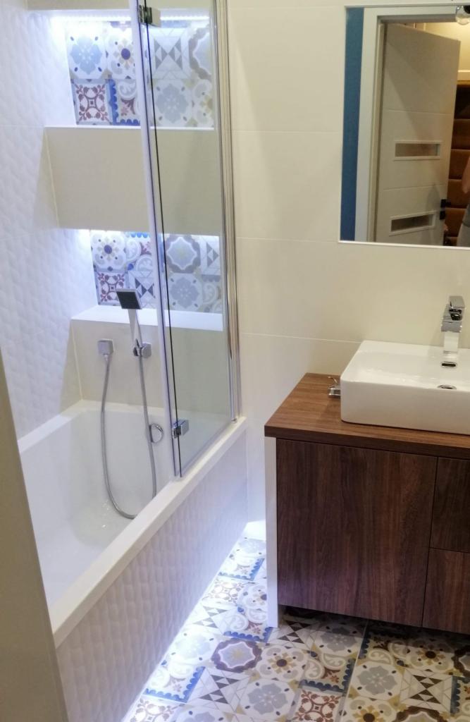 Wykończona łazienka z wanną za szklaną ścianką, umywalką na szafce z drewna i kolorową mozaiką na podłodze. Wnęki z podświetleniem eksponują dekoracyjne płytki.