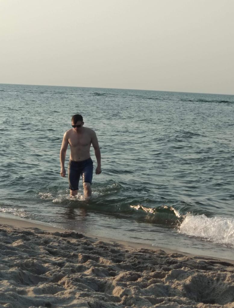 Mężczyzna w okularach przeciwsłonecznych wchodzi do morza, woda sięga mu do kolan, widoczna piaszczysta plaża i delikatne fale.