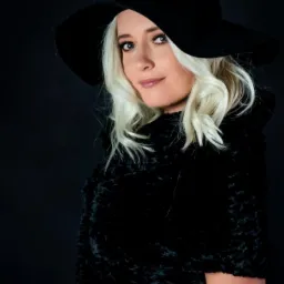 Portret kobiety z blond lokami, w czarnym kapeluszu z szerokim rondem i czarnym futrze na ciemnym tle. Ujęcie w lekkim półprofilu, z delikatnym uśmiechem.