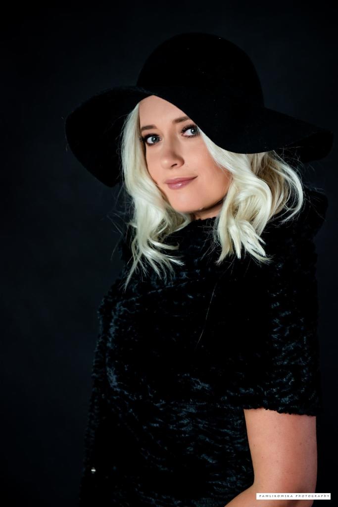 Portret kobiety z blond lokami, w czarnym kapeluszu z szerokim rondem i czarnym futrze na ciemnym tle. Ujęcie w lekkim półprofilu, z delikatnym uśmiechem.