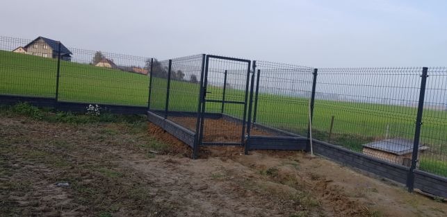Ogrodzenie panelowe z bramką, z fundamentem z desek kompozytowych, na nierównym terenie, w tle dom jednorodzinny.